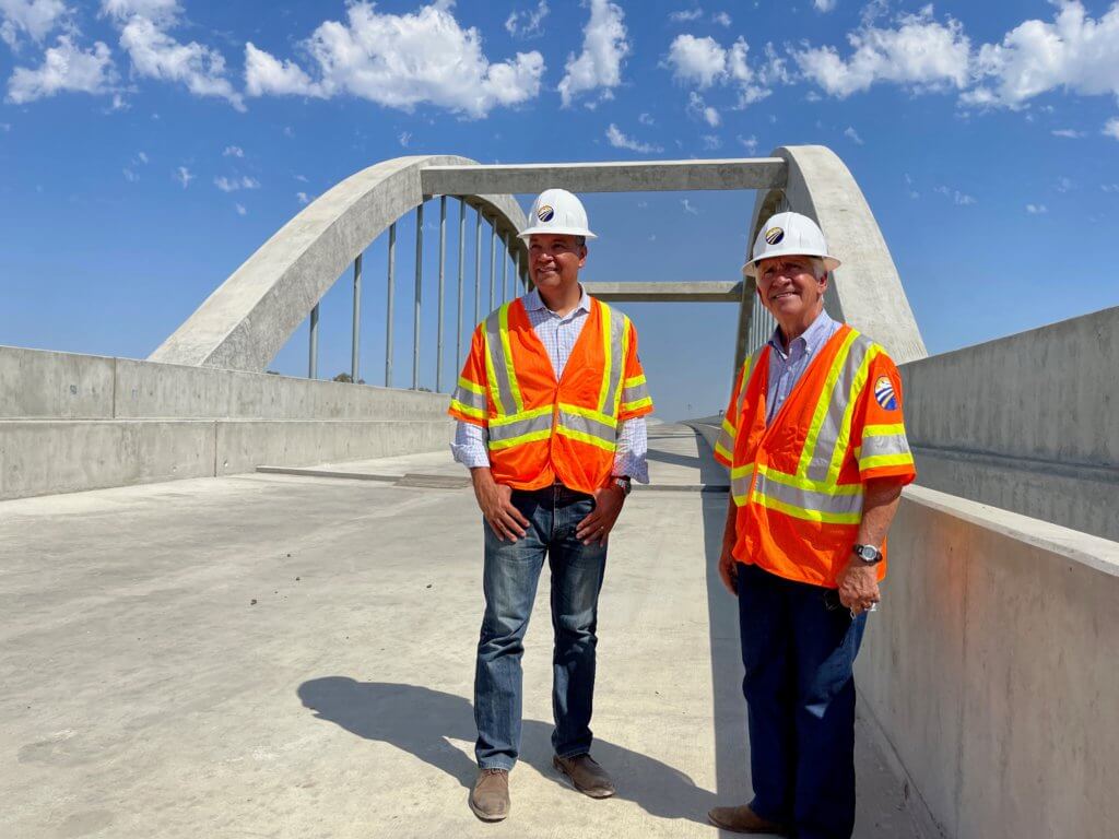 California Senator Alex Padilla and Congressman Jim Costa stand on the award winning San Joaquin River Viaduct with the singature arches and a beautiful blue and cloud flecked sky behind them.