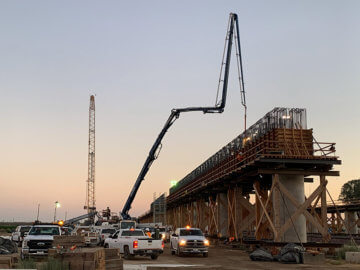Viaduct structure under construction at dusk with pickup trucks parked on left side and cement truck piping cement to top of structure