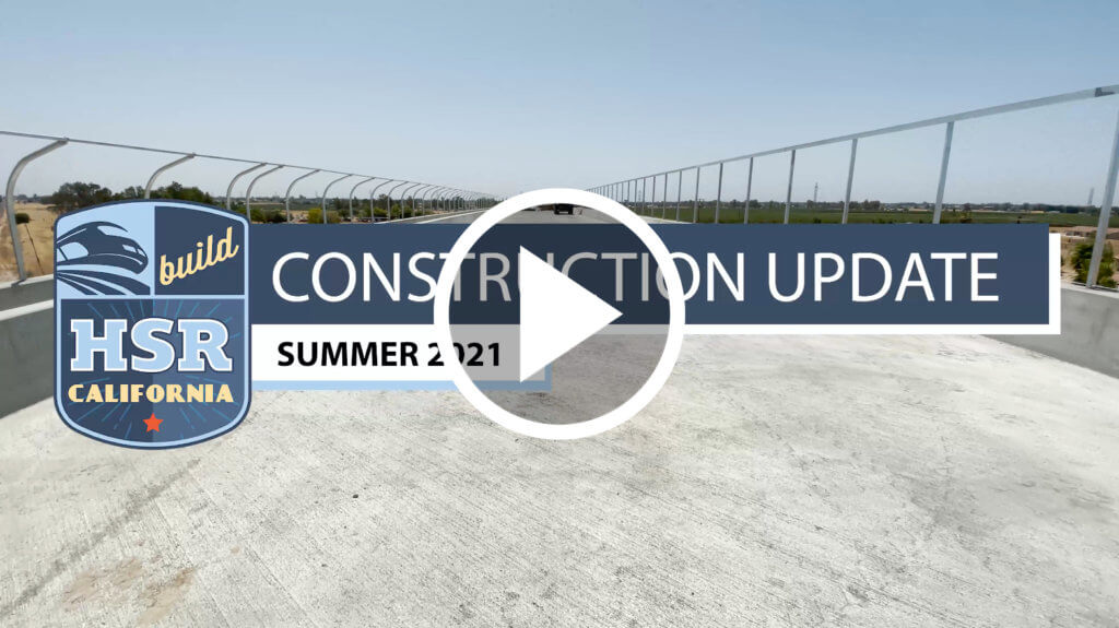 A still of the video, which reads Construction Update Summer 2021, shwoing a complete concrete road deck over tracks, and a super imposed play button, encouraging seeing readers to click the image to be directed to the video on YouTube.