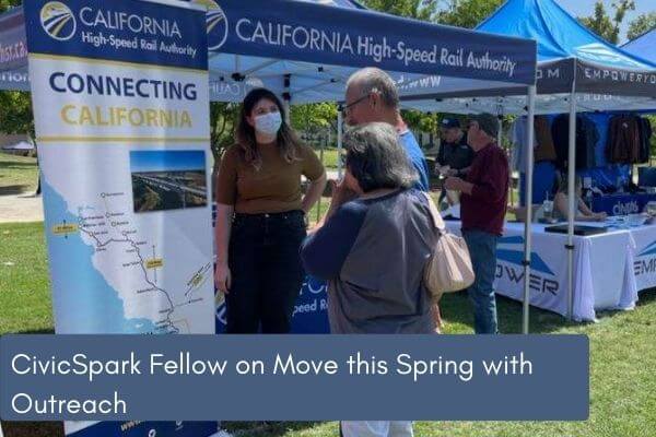 Women stands in front of an outreach pop-up banner with a California high-speed rail alignment map that shows a statewide angle. Woman talking to two community members.