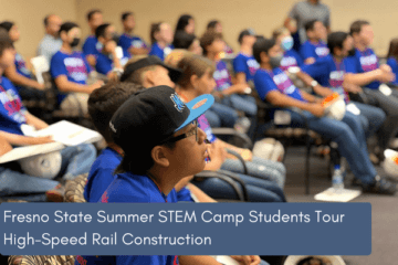 A student looks intently to the front of the room during a presentation. Student is sitting with a larger group of students and is wearing a hat. 