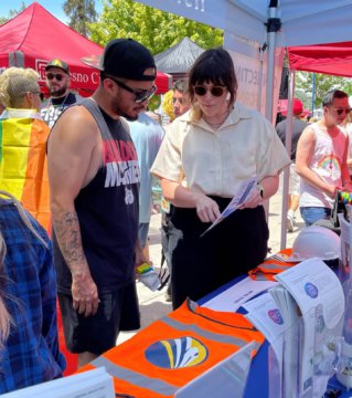 Photo of a women tabling at a fair speaking to a member of the public while pointing at a paper.