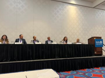 Panel discussion with many people on stage sitting behind mics. 