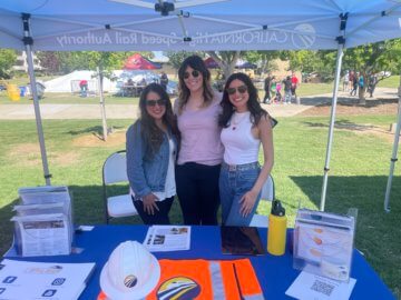 three women smiling and tabling at a public fair. Title of the article included in the graphic. 