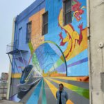 Wide shot of mural in Fresno’s Chinatown, with artist Mauro Carrera in front. Painting includes the conceptual rendering of CA HSR trains, an outline of California and her counties, a traditional Chinese dragon, and lines drawing the viewer toward the horizon and conceptually the future. A more detailed description of this image is available upon request to the email address info@hsr.ca.gov