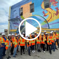 Photo of large group of people in construction vests and hard hats in front of a mural. Photo has a play button on it. 