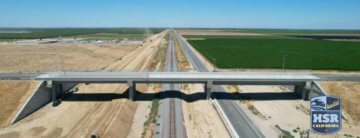 Aerial view of an overpass that goes over a road, train tracks and a dirt path that is the future alignment of high-speed rail. Photo has a BuildHSR logo on the bottom corner.