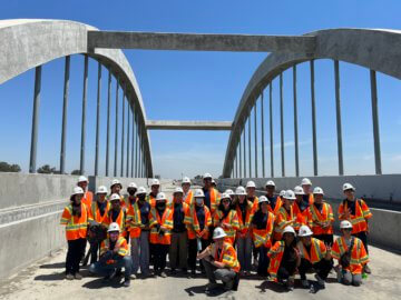 Group of high school students on a bridge with a large arch over the group. All students, about 30 of them, in construction hard hats and bright safety vests. 